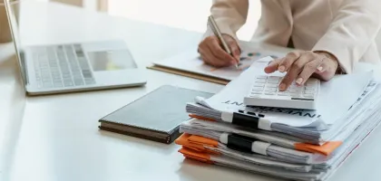 Photo of a woman using a calculator with documents and a laptop